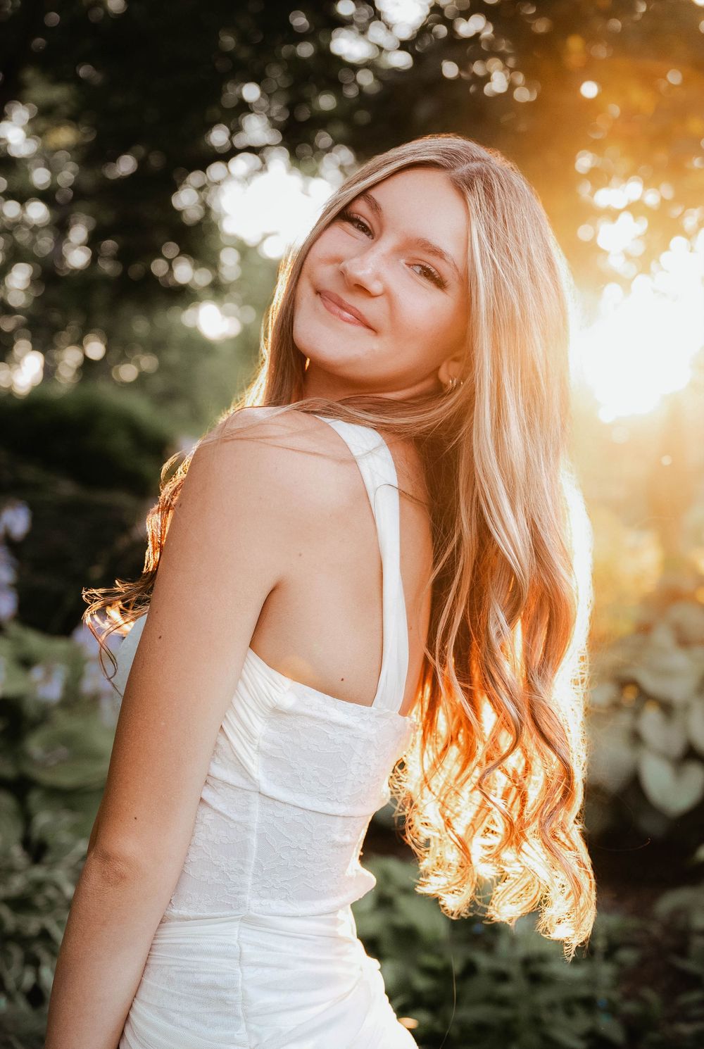 Young woman with long blonde hair smiling in golden sunset light, wearing a white dress in a lush garden setting.