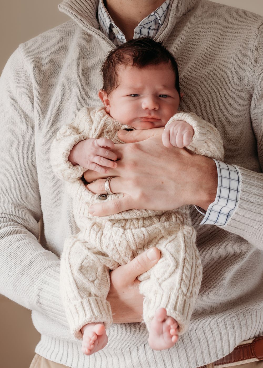 A parent in a beige knit sweater cradles a sleeping newborn baby dressed in a cream cable-knit outfit.