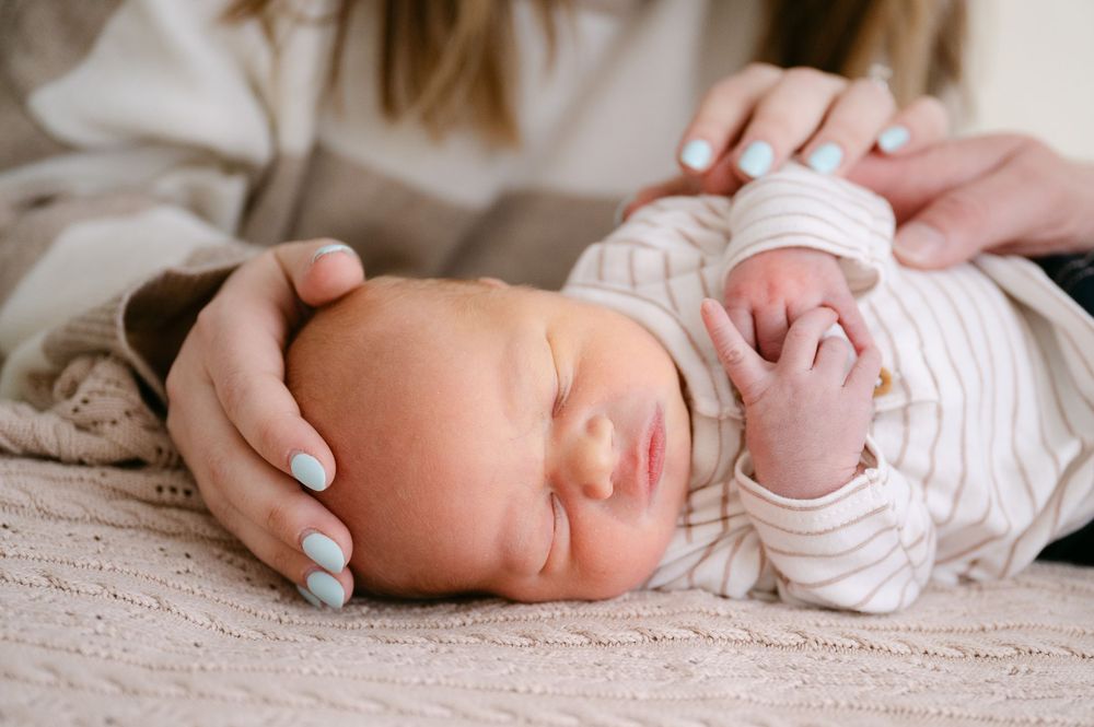 Newborn baby sleeping peacefully while mother with blue nails gently cradles their head on a soft blanket.