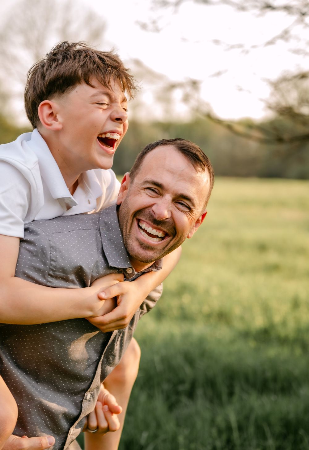 A joyful father gives his laughing son a piggyback ride outdoors in a sunny green field.