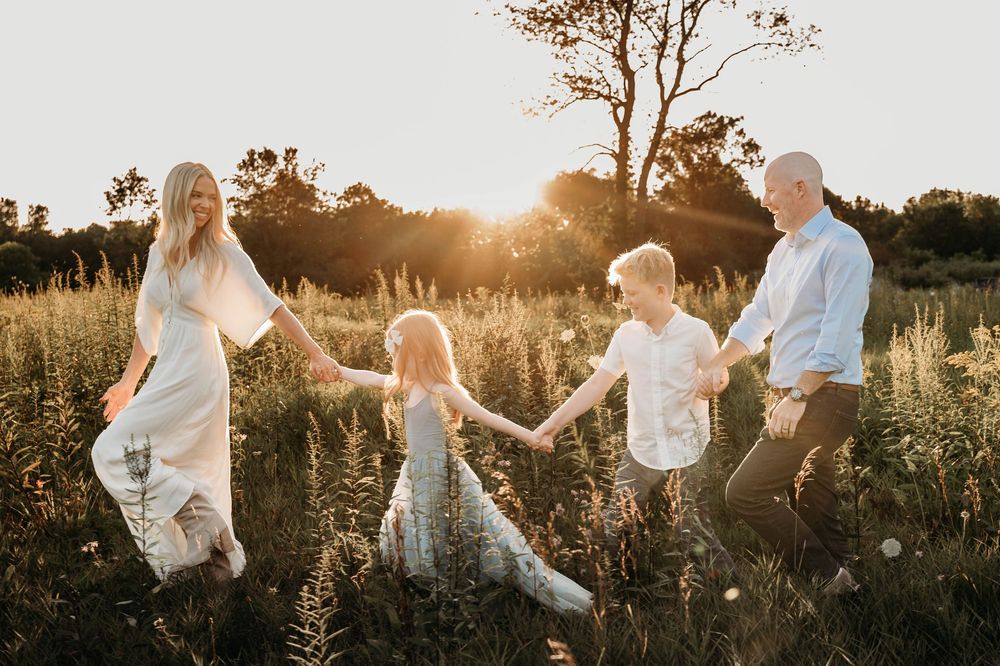Family of four holding hands in a sunlit meadow at golden hour, surrounded by wildflowers and trees.