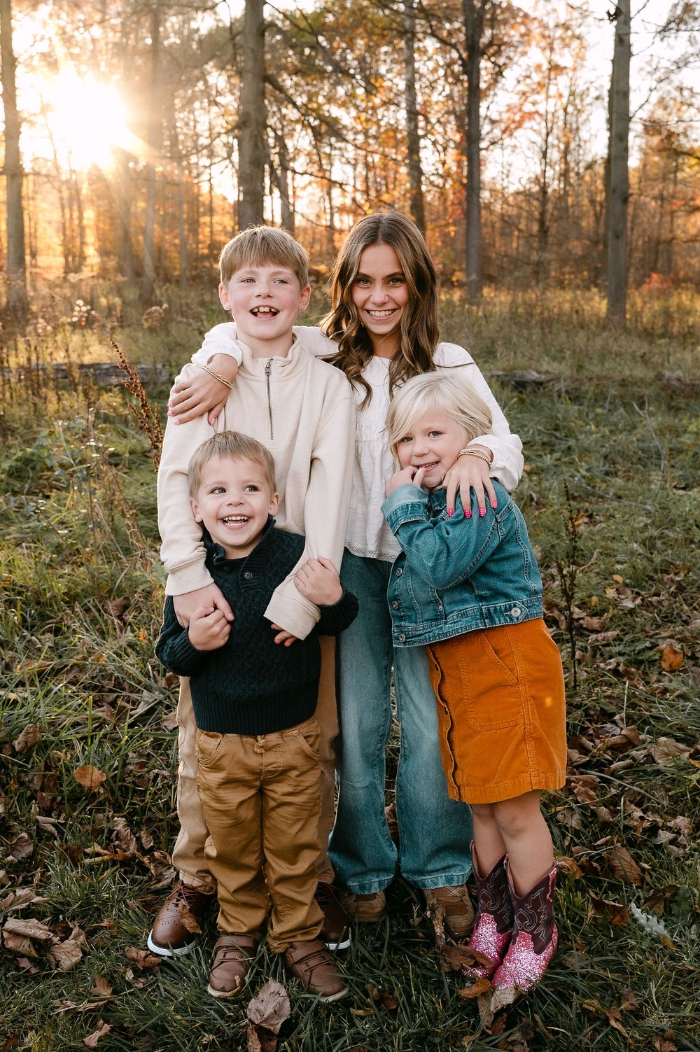 A woman and three young children smile together in a sunlit autumn forest during a family portrait session.