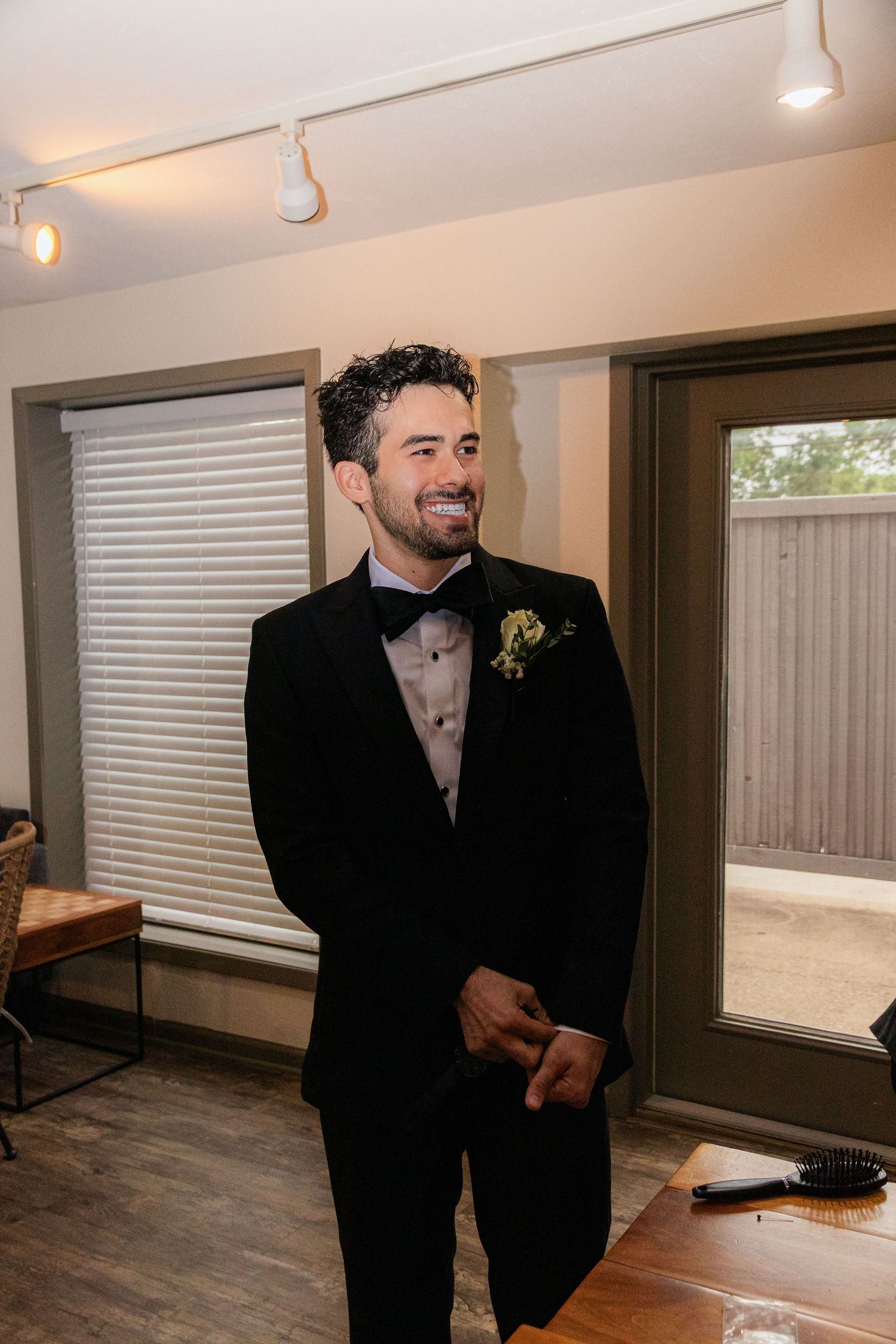 Smiling groom in black tuxedo with boutonniere standing indoors on wedding day.