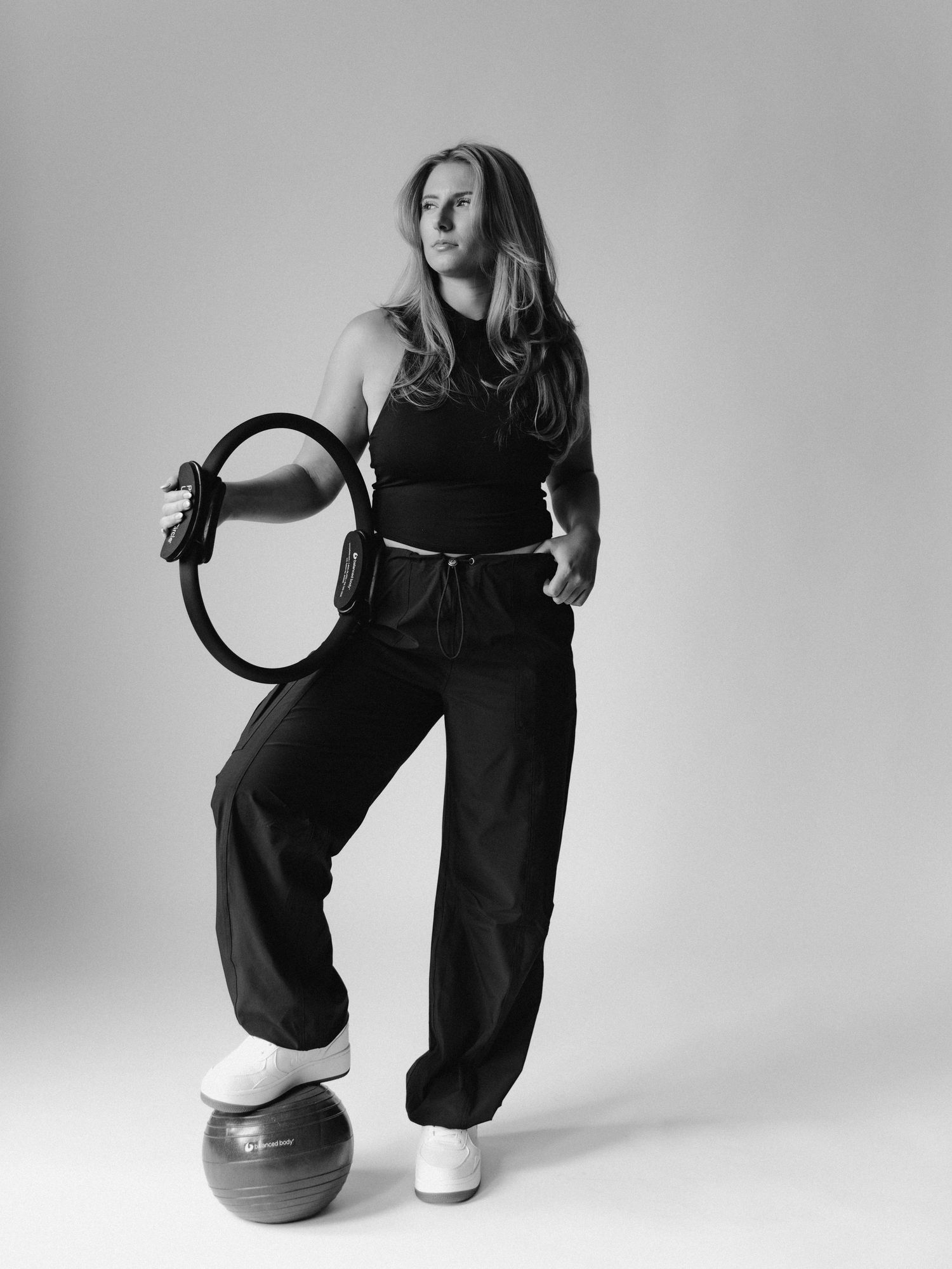 Black and white photo of a woman in athletic wear holding a pilates ring with a fitness ball underfoot.