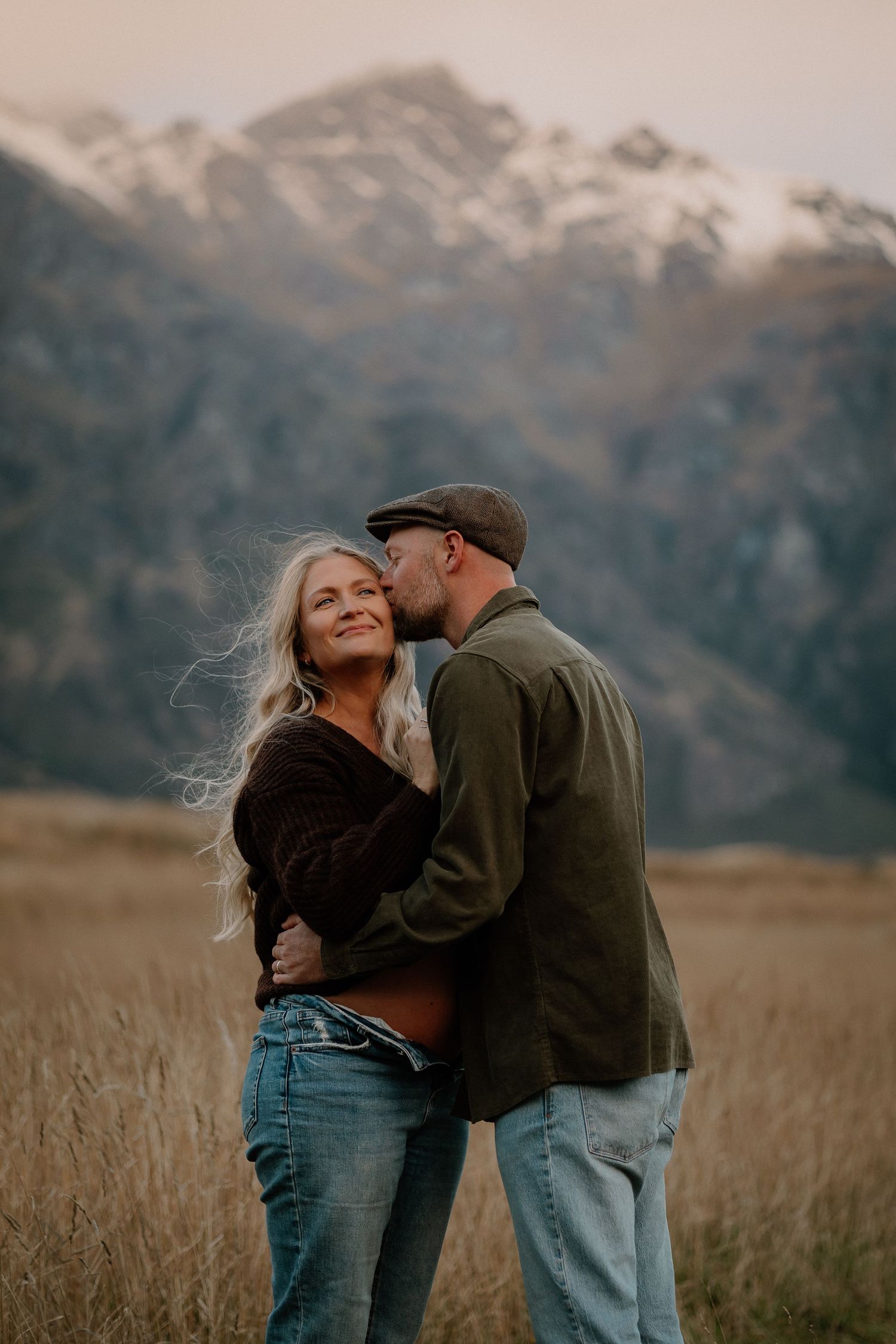 A couple embraces romantically in a golden field with snow-capped mountains backdrop during golden hour.