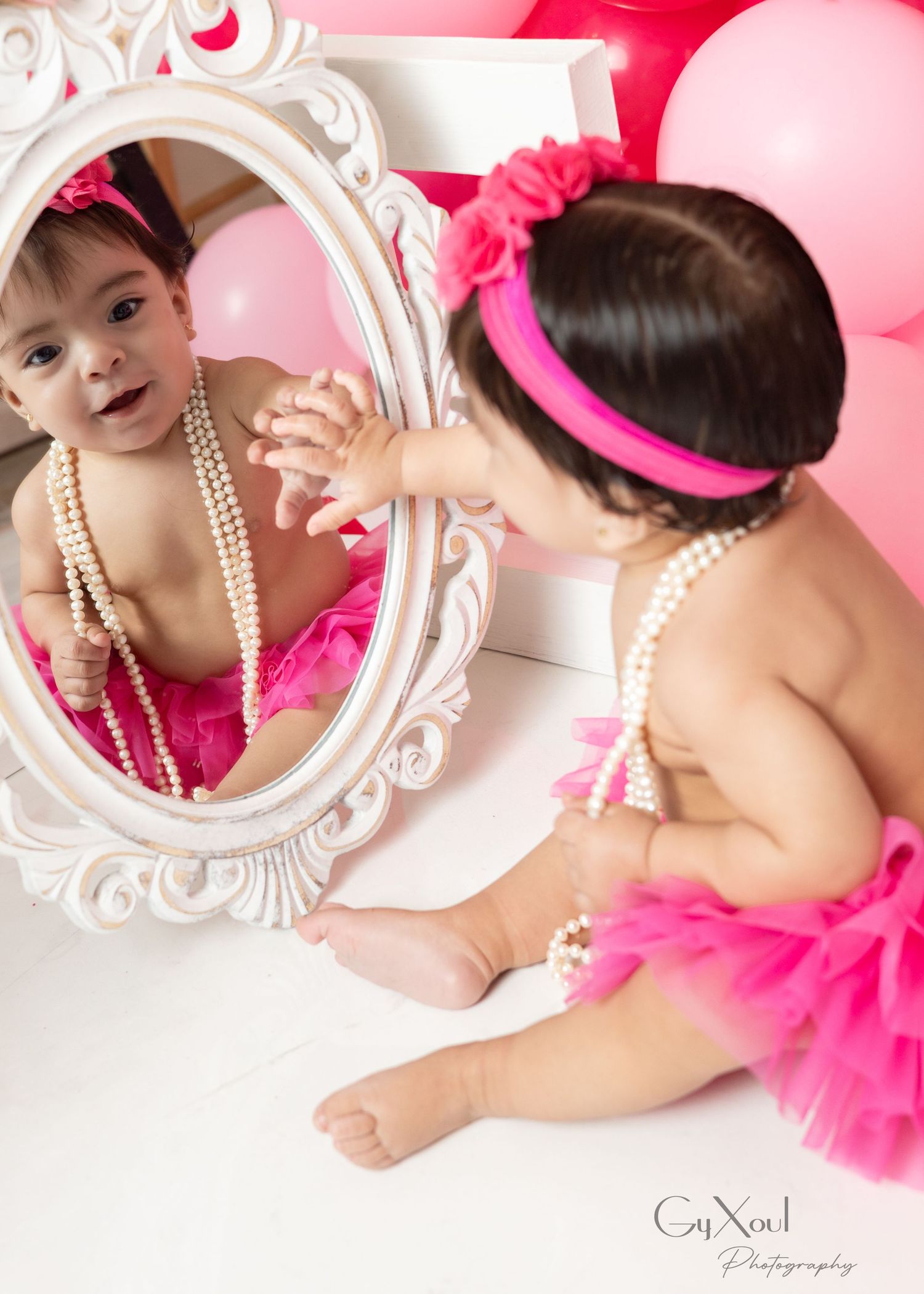 Baby girl in pink tutu and pearl necklace touches her reflection in an ornate white mirror, surrounded by pink balloons.