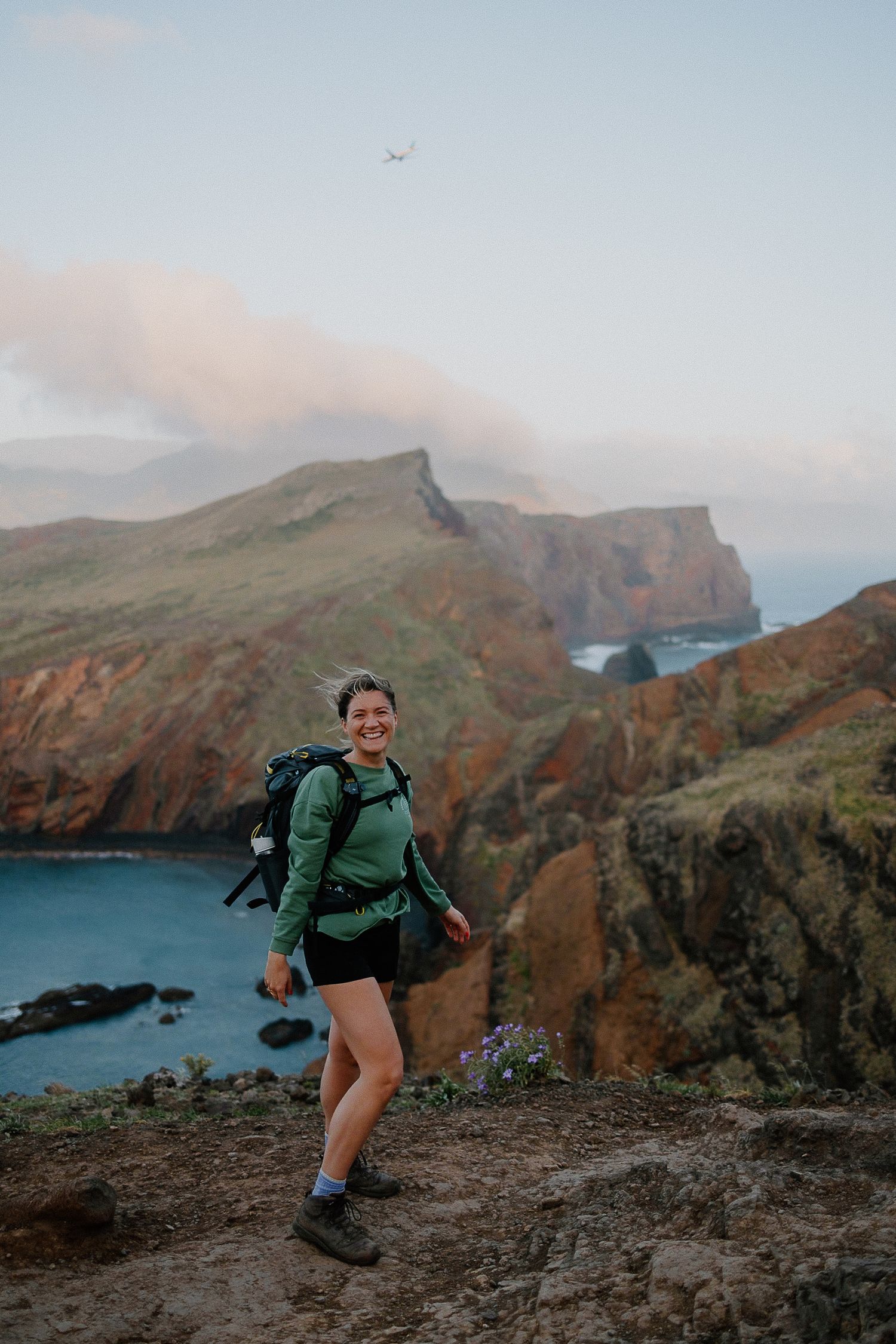A smiling female hiker with backpack stands on a scenic coastal cliff trail with dramatic mountains and ocean views.