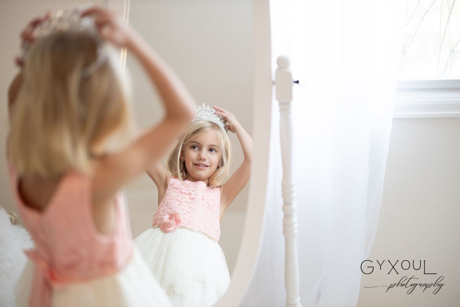 Young flower girl in pink and white dress adjusts tiara while smiling at her reflection in a tall mirror.