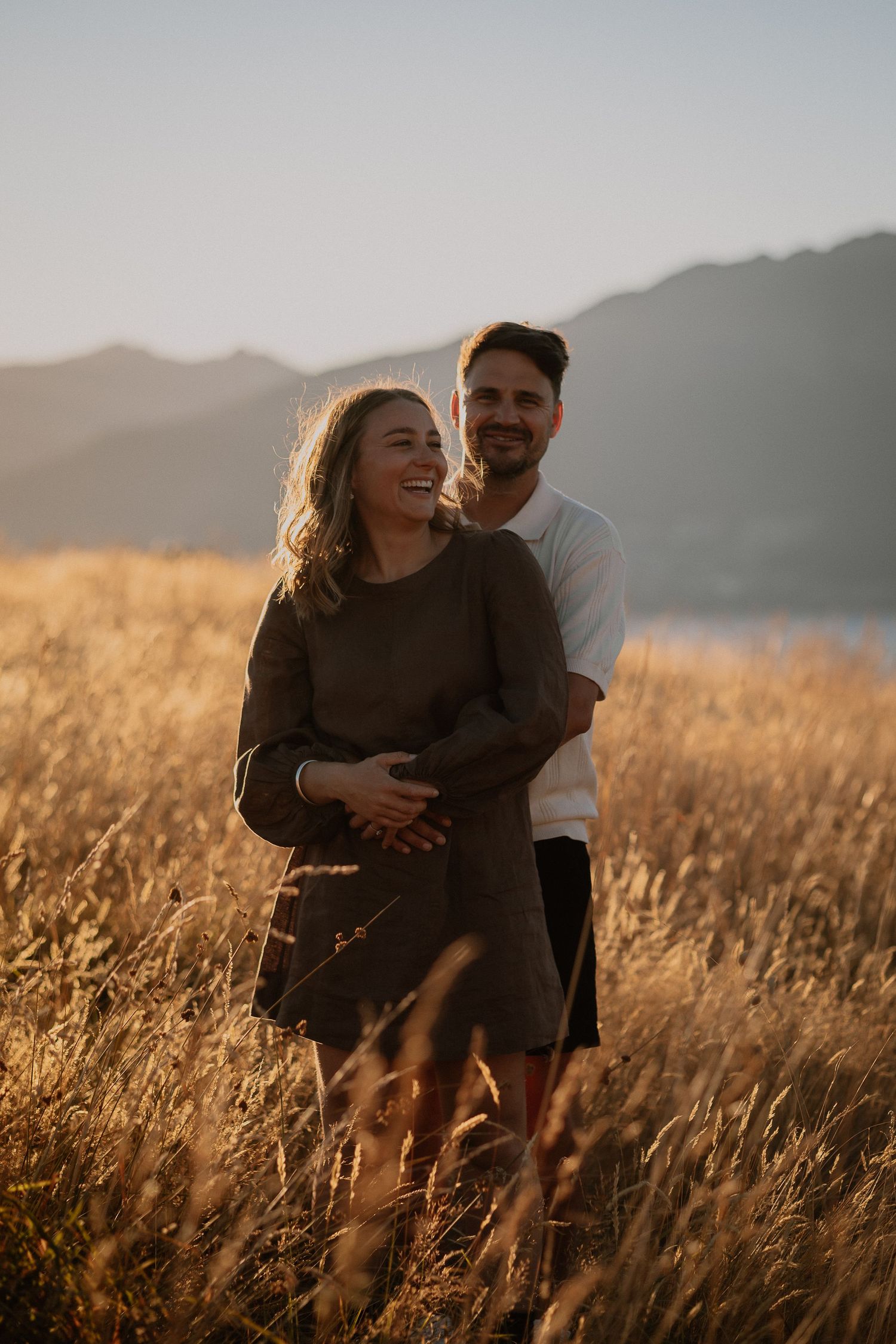 A couple embraces in a golden field at sunset, with mountains and a lake in the background.