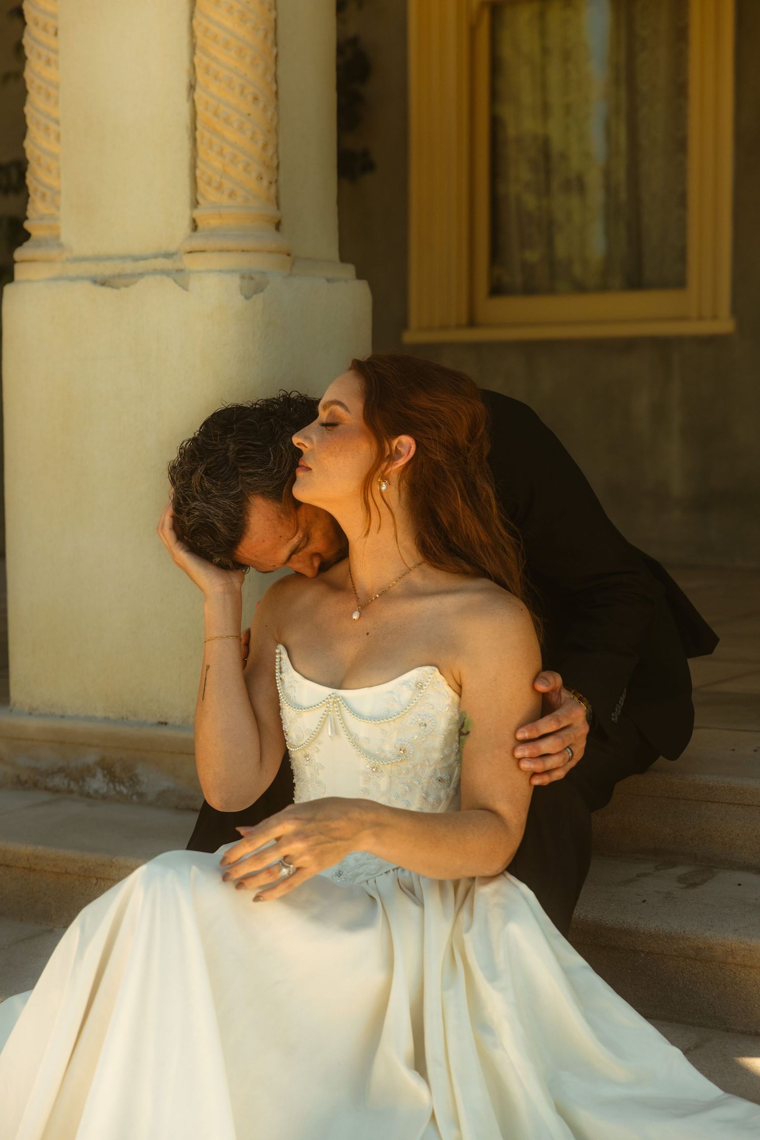 A groom in a dark suit kisses a bride in a white strapless gown on the steps of the Kimberly Crest House wedding venue.