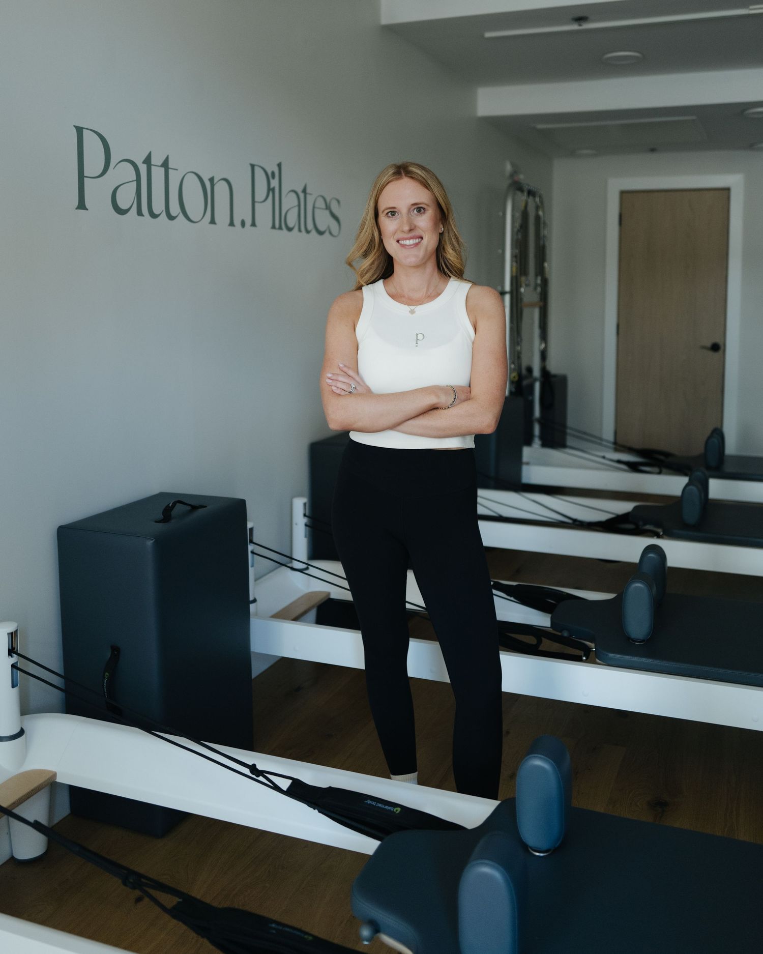 Woman in white top and black leggings poses confidently in Patton Pilates studio with reformer equipment.