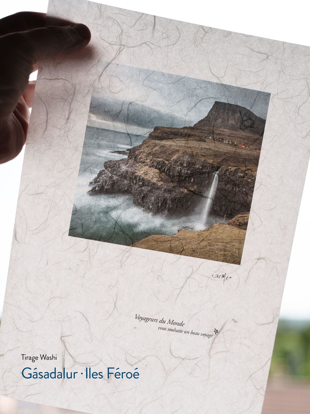 Hand holding a Washi print of Gásadalur waterfall in Faroe Islands, featuring dramatic cliffs and misty ocean waves.