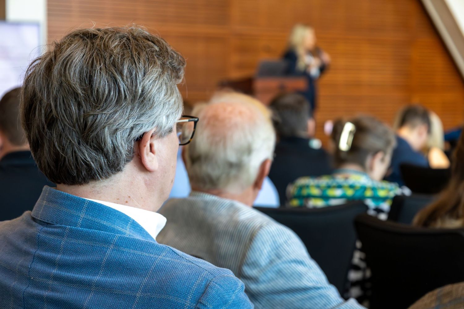 Audience members seated at a professional conference listening to a speaker presenting at the front of the room.