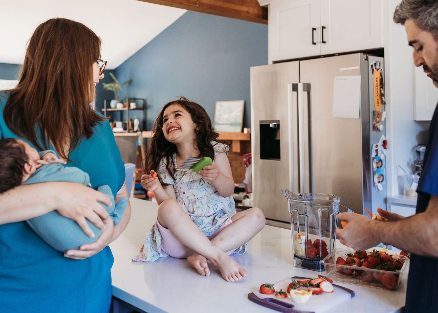 Family enjoying time in a bright kitchen, mom holding baby, daughter sitting on counter, dad preparing fresh fruit smoothie.