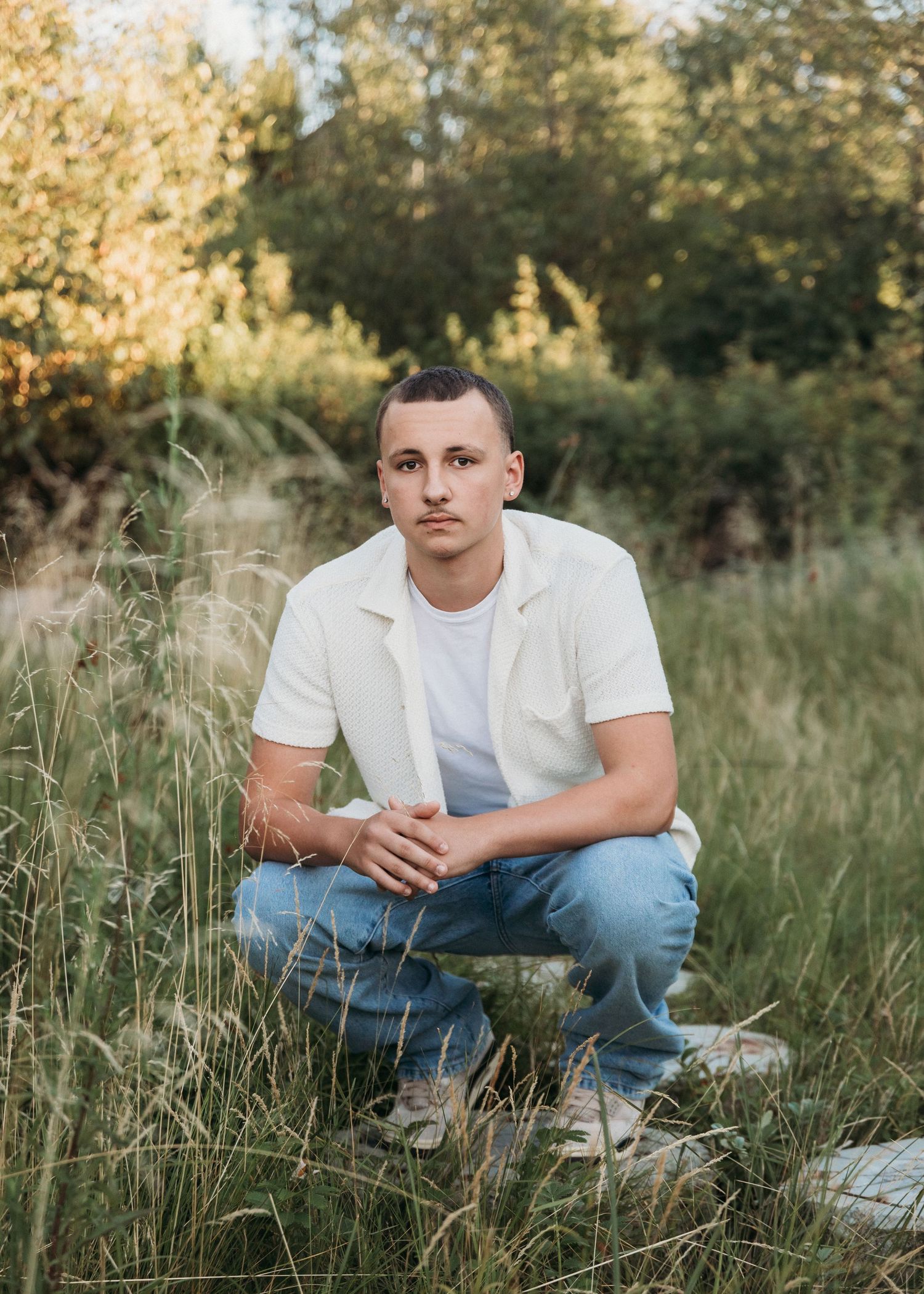 Young man in white shirt and jeans crouching in tall grass with green trees in golden hour light.