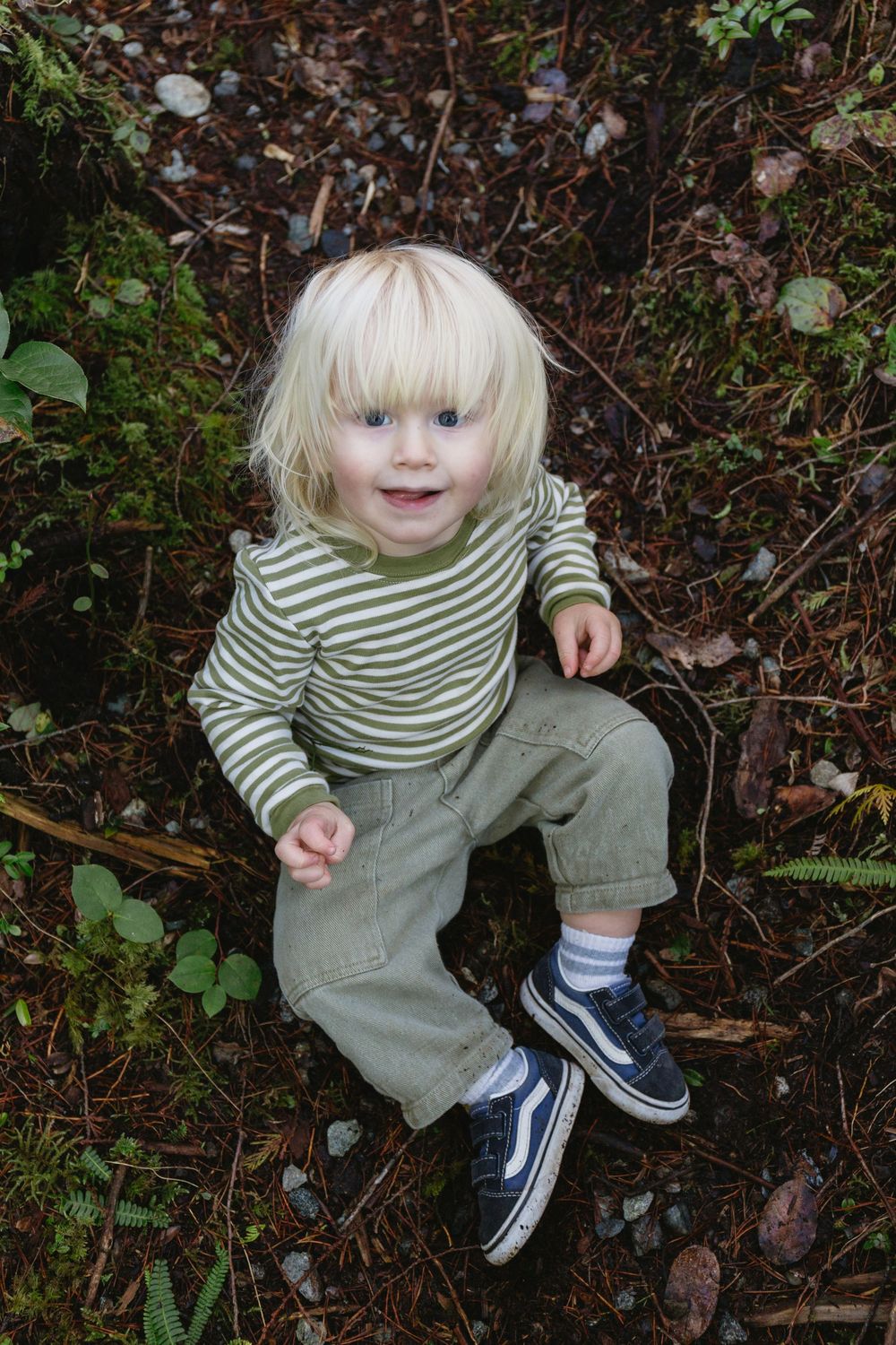A smiling blonde toddler in a striped shirt sits on a forest floor looking up.