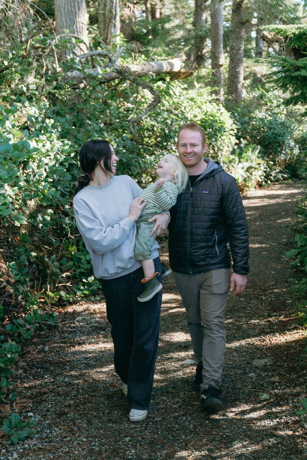 Happy family with toddler walking on a lush forest trail, mother carrying child, father smiling.