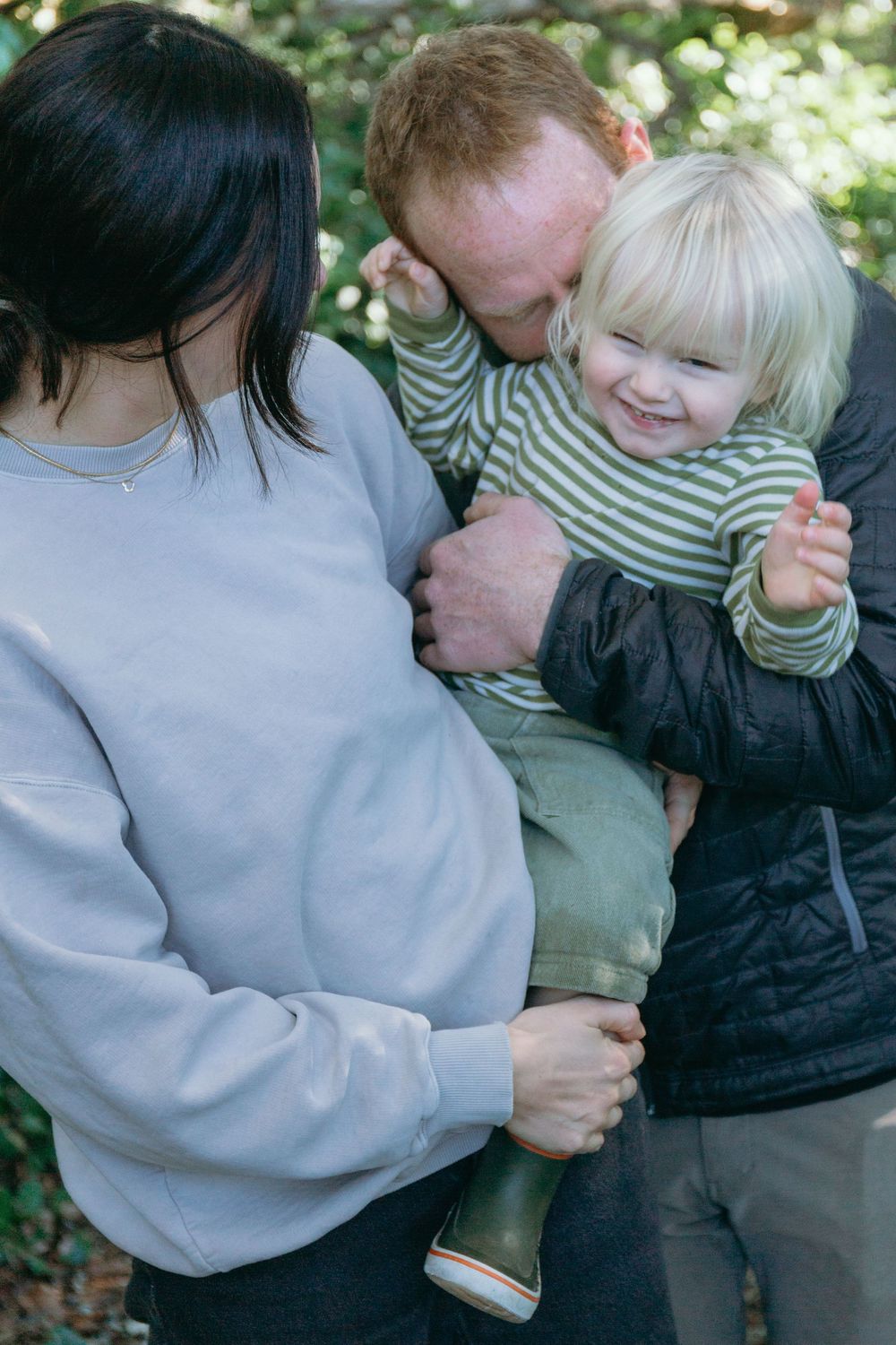 Father kissing smiling blonde toddler in striped shirt while mother holds child in forest setting.