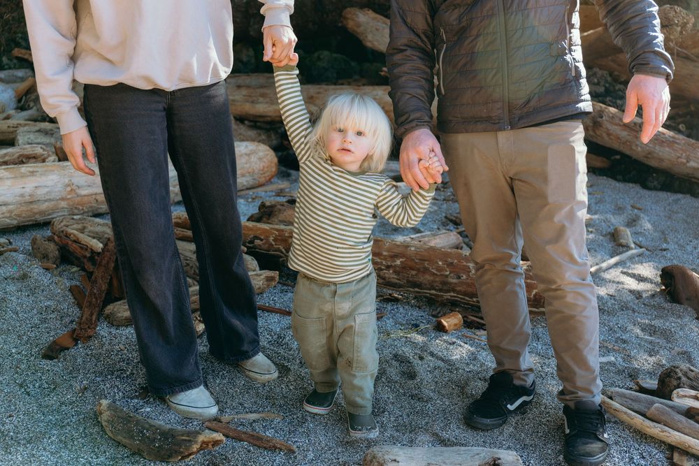 Young blonde toddler in striped shirt holding parents hands on sandy beach with driftwood.