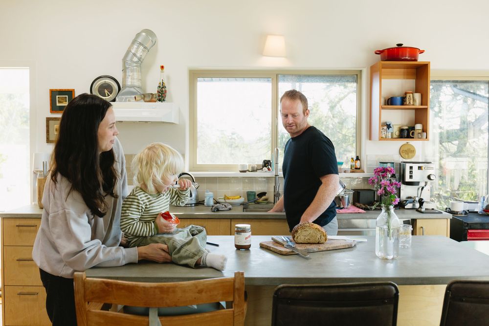 A family gathers in a bright modern kitchen as a father slices bread at the counter.