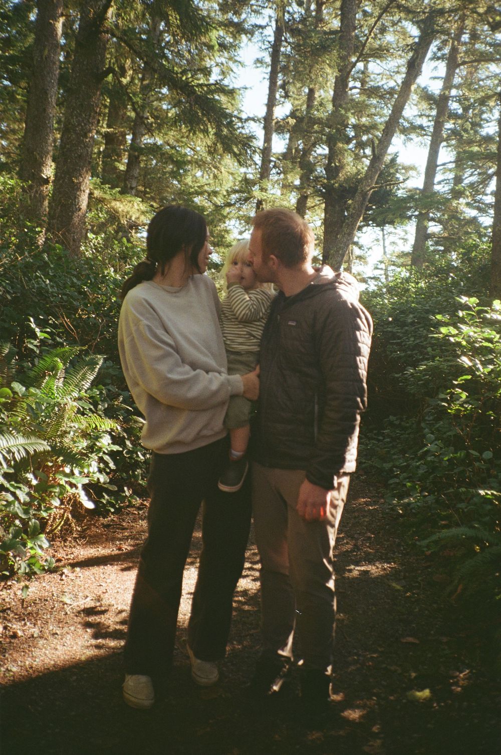 A couple with dark hair shares a tender moment with their blonde toddler on a forest path.