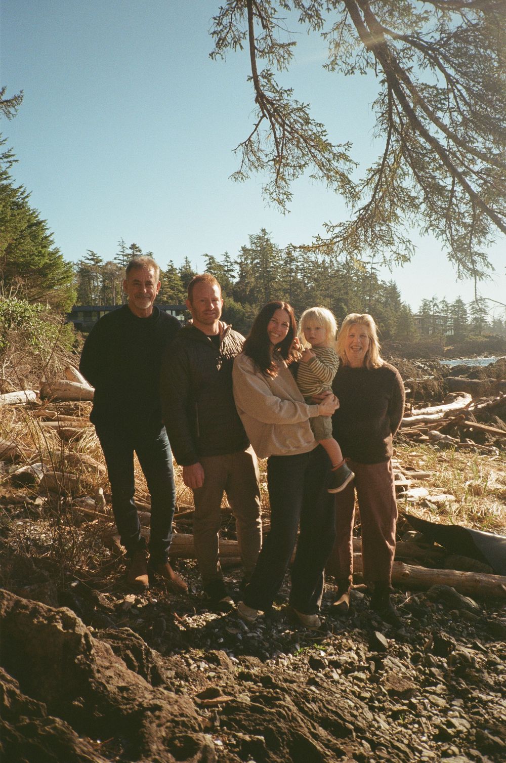 A family of five poses together on a rocky beach surrounded by driftwood and pine trees.