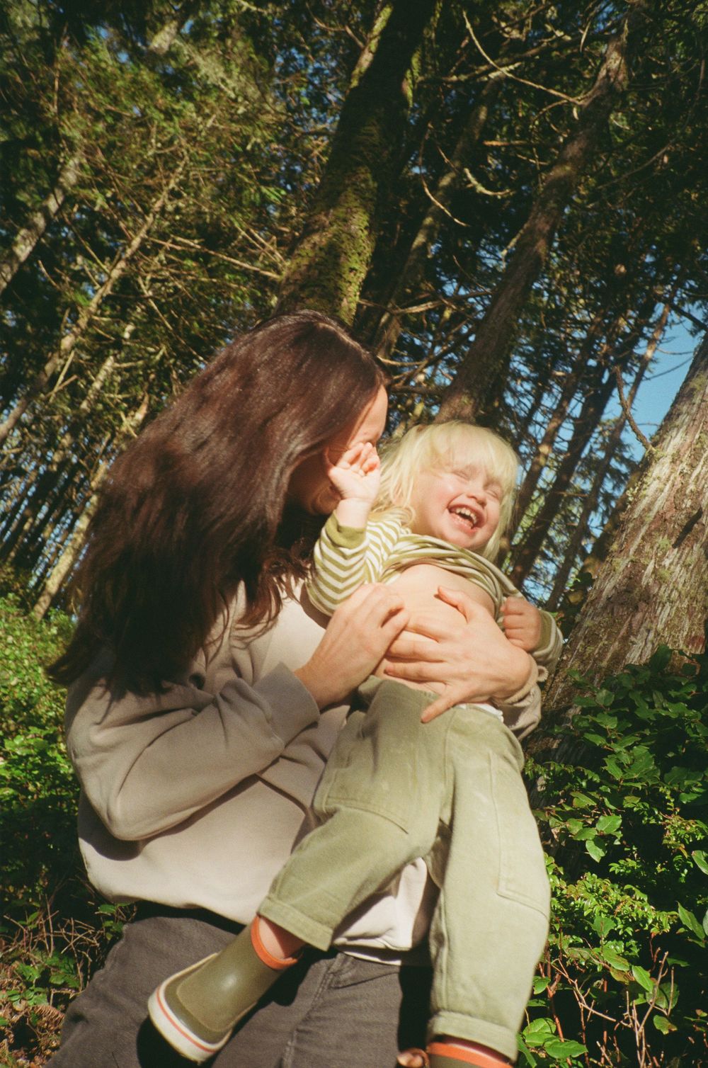 A dark-haired mother lifts her laughing blonde toddler in a sunlit forest.