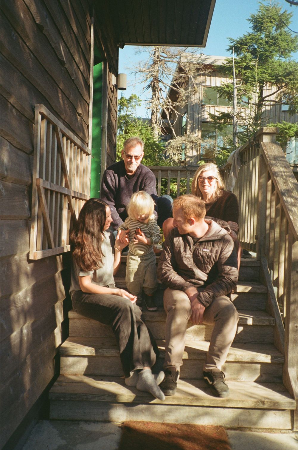 A multigenerational family of five sits together on wooden outdoor stairs in warm sunlight.