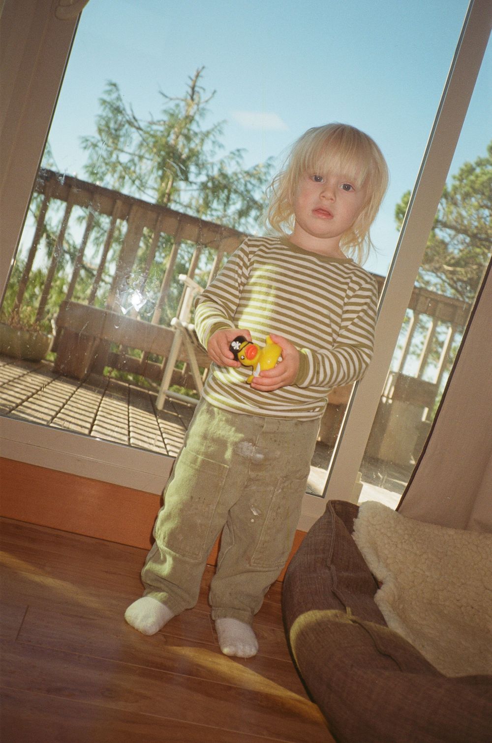 A blonde toddler in a striped shirt holds a rubber duck toy while standing by a glass door.