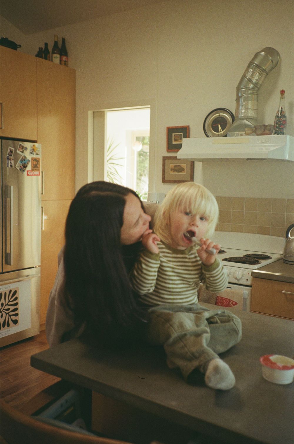 A mother kisses her blonde toddler sitting on a kitchen counter holding a spoon.