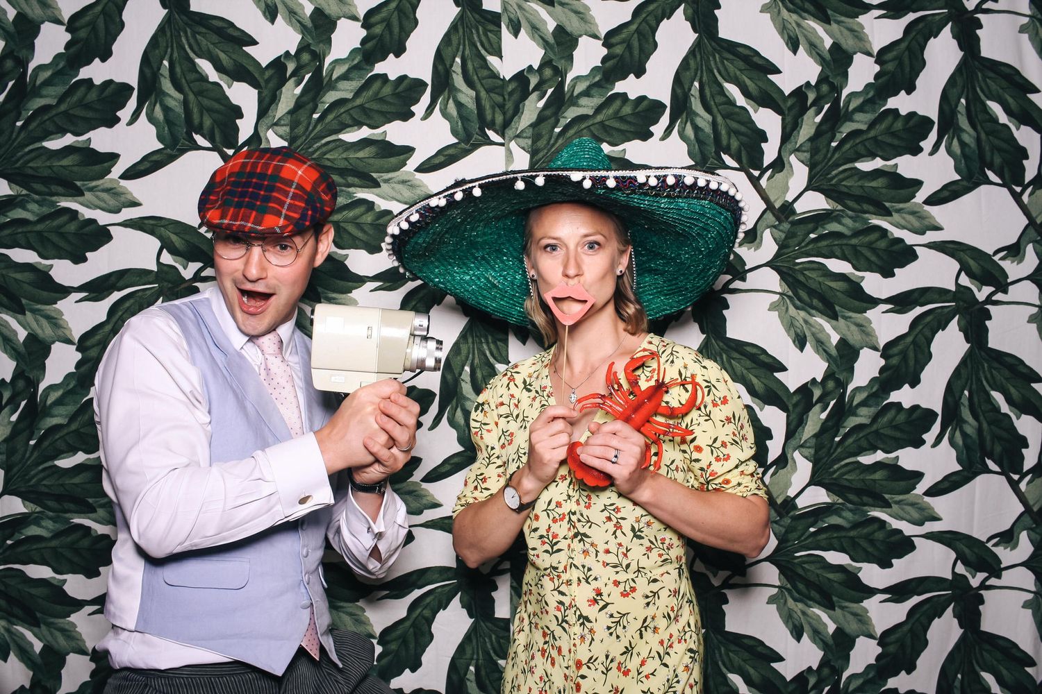 Two people in festive costumes pose in a photo booth with tropical leaf wallpaper backdrop.