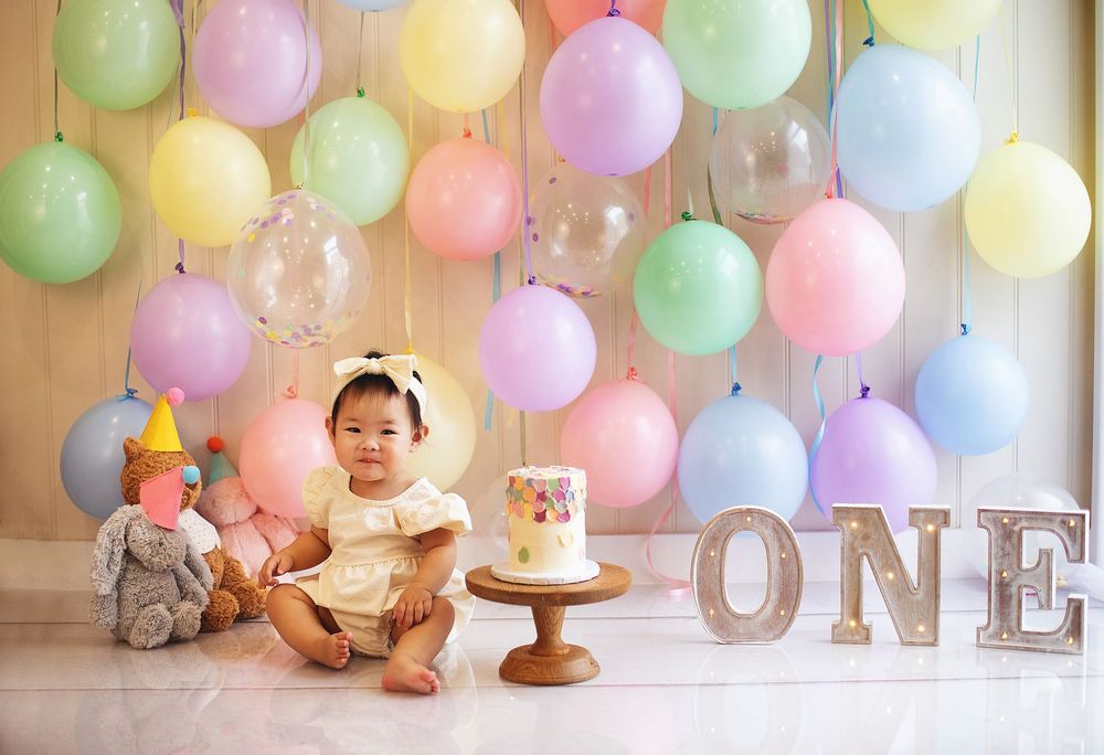 Baby girl in yellow dress sits before a colorful balloon backdrop with ONE letters at her first birthday cake smash.