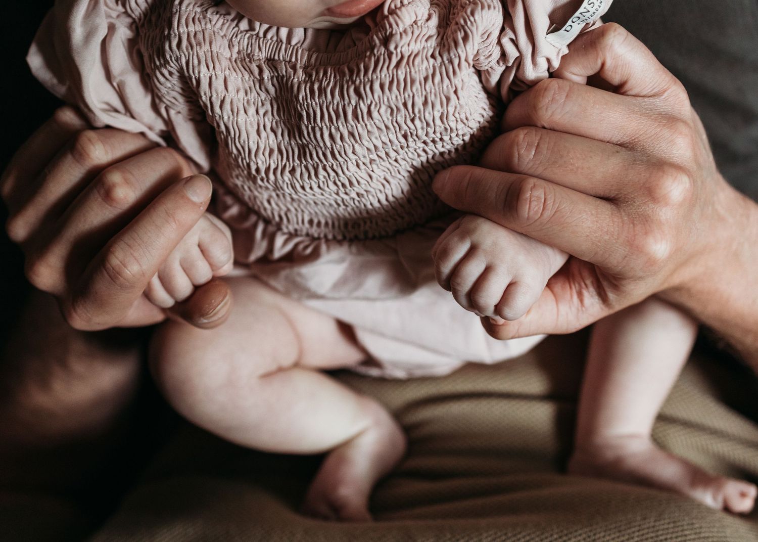 Close-up of a newborn baby held by adult hands, wearing a pink smocked dress with tiny bare feet visible.