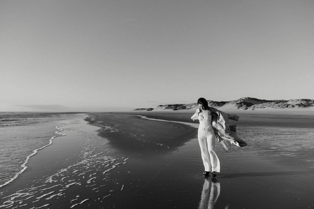 Black and white photo of a woman in white dress dancing freely on a serene beach with waves and sand dunes.