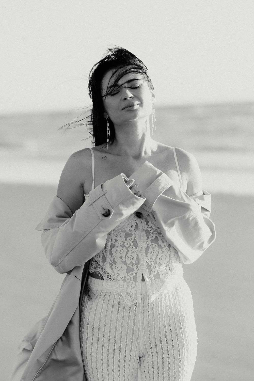 Woman in lace bodysuit and trench coat poses on beach with eyes closed in black and white.