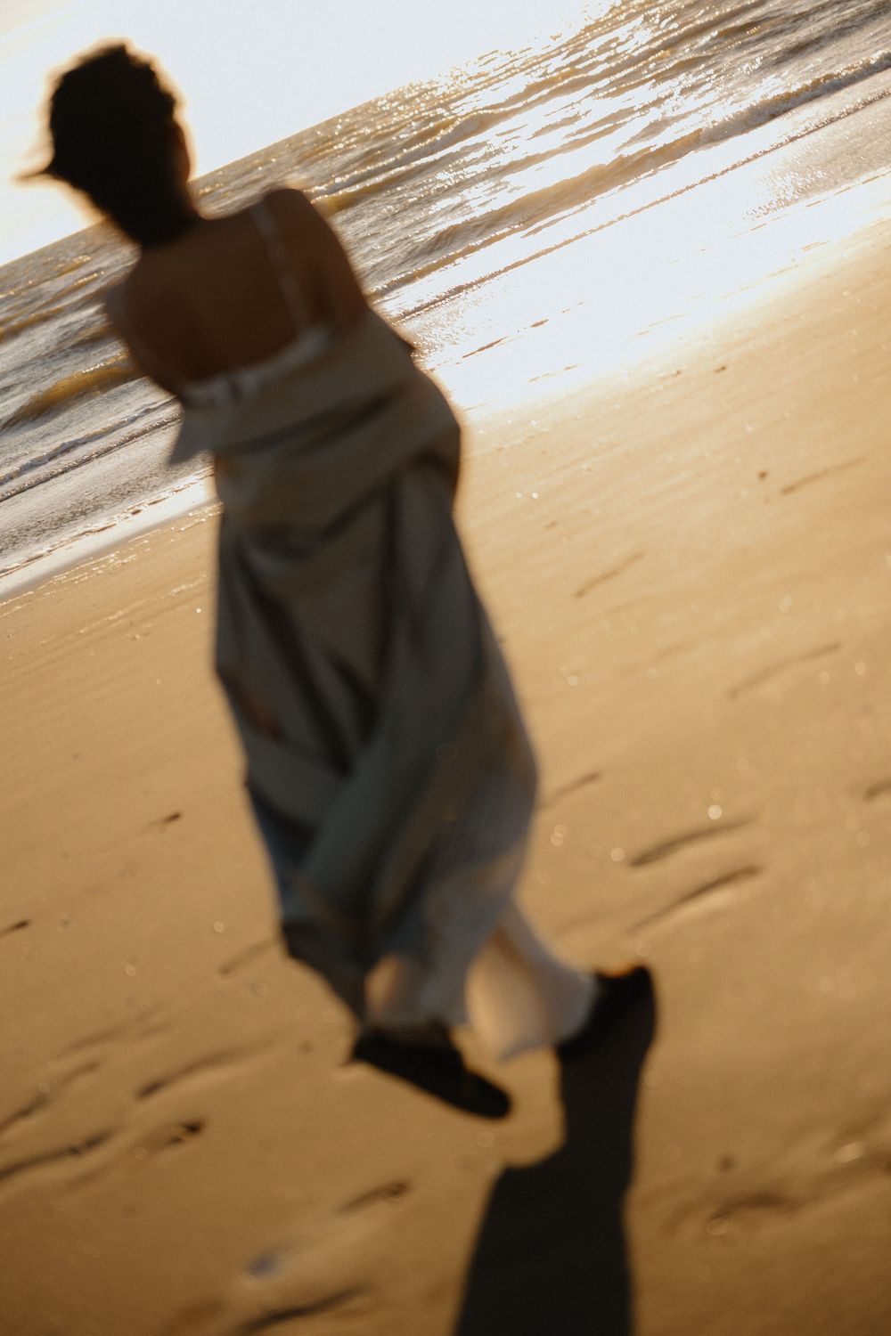 Blurred silhouette of woman in white dress walking toward ocean on sandy beach at sunset.