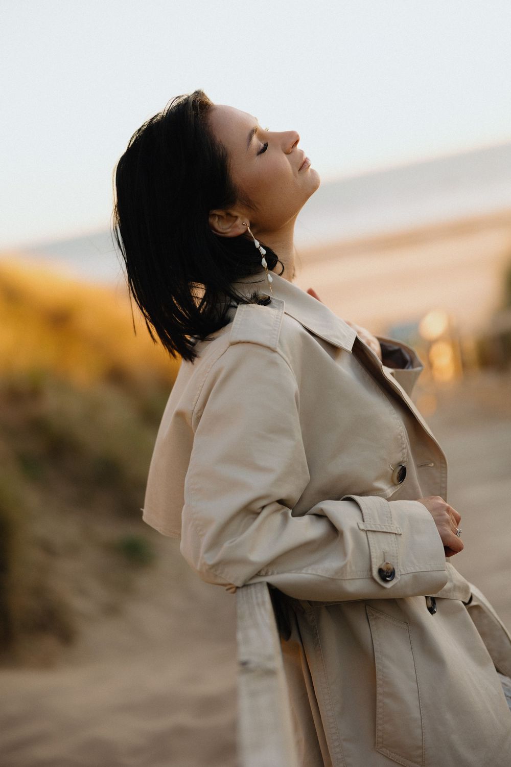 Woman in beige trench coat with eyes closed, enjoying fresh air outdoors at golden hour.