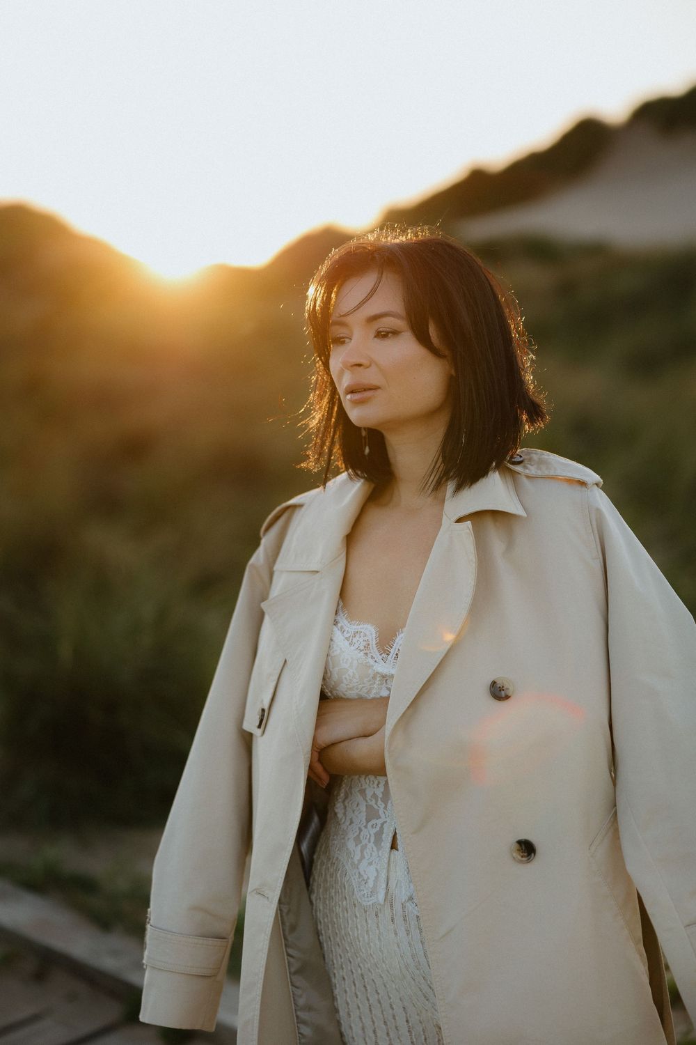 Woman in beige trench coat standing outdoors at golden sunset with hills in background.