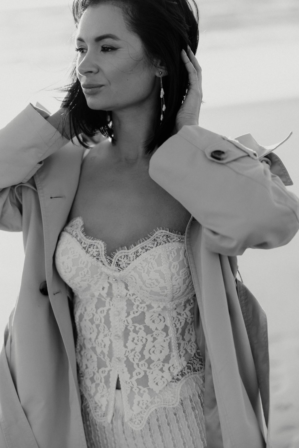 Black and white portrait of brunette woman in lace dress and oversized coat posing outdoors near the sea.