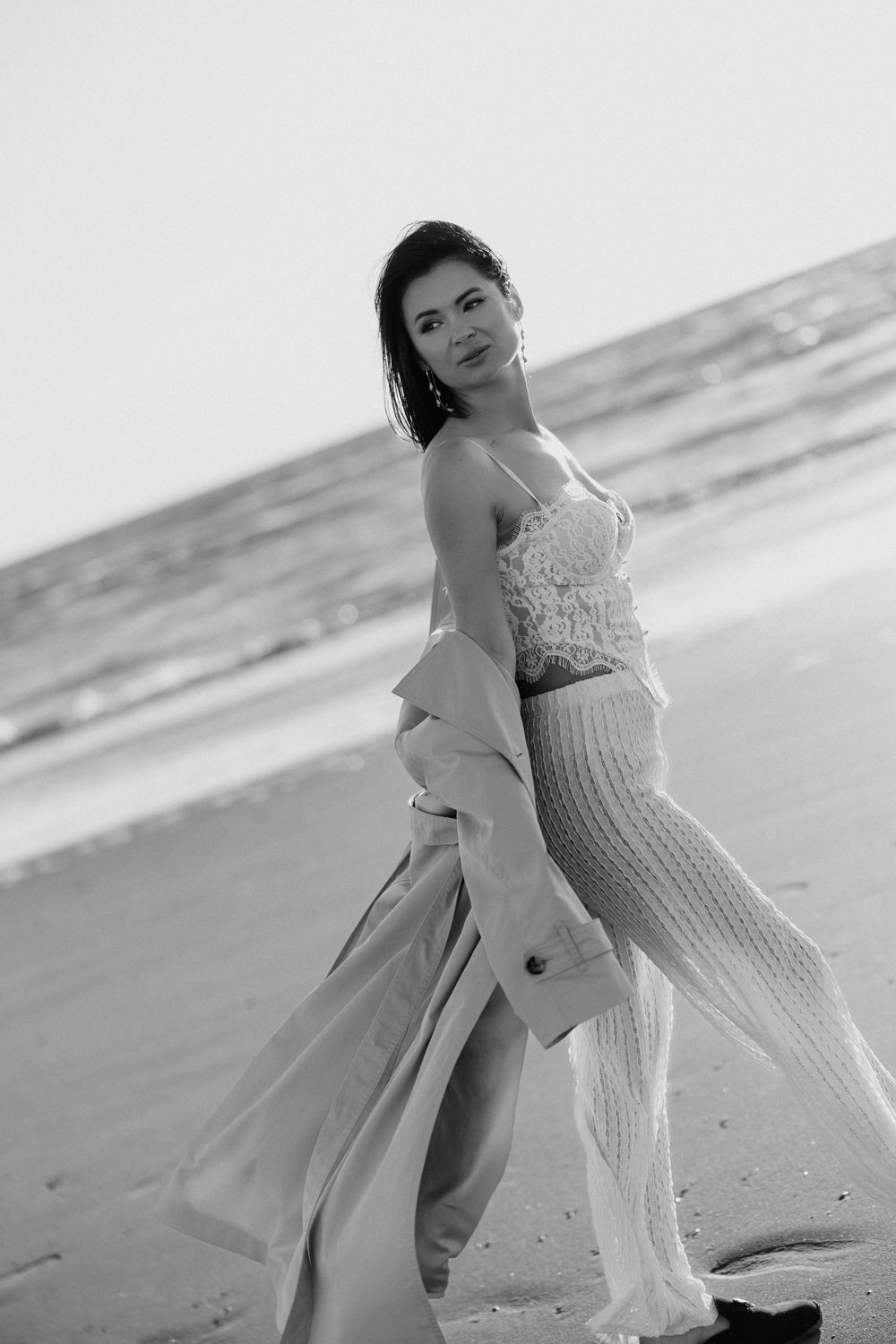 Woman in lace top and knit pants walks on beach holding trench coat in black and white photo.