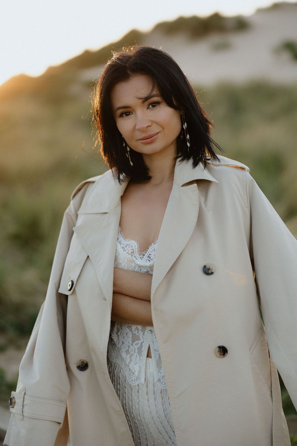 Woman in beige trench coat and white lace outfit poses outdoors at golden hour with sandy dunes backdrop.