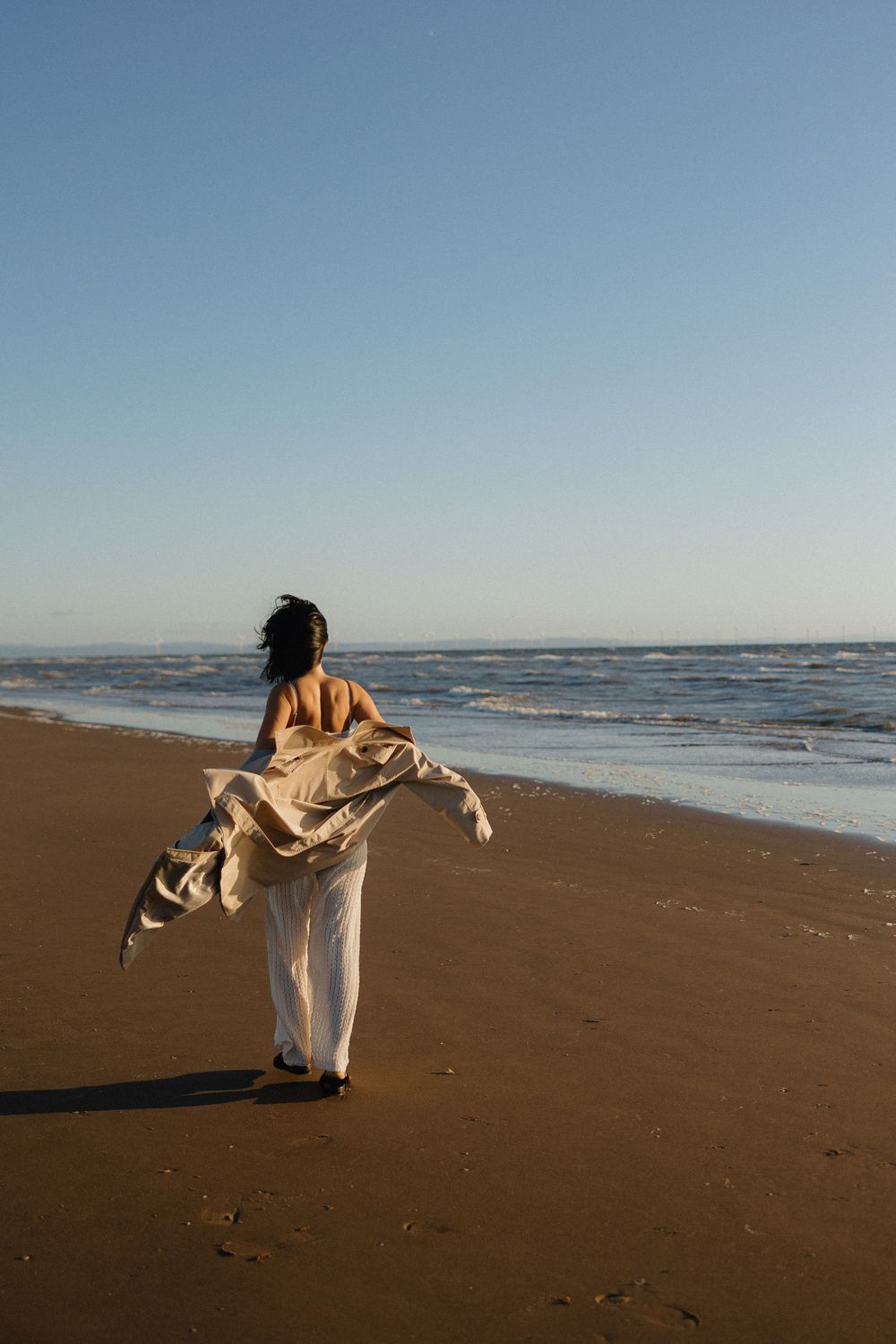 Woman in white pants and flowing jacket walking along sandy beach shore at golden hour with calm ocean waves.