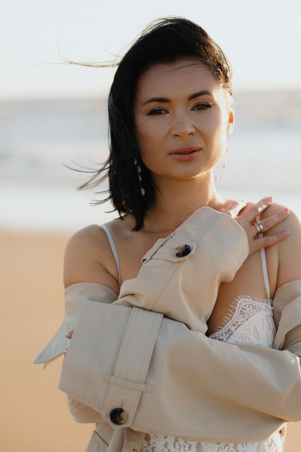 Woman in beige jacket and white lace top poses on sandy beach with ocean in background.