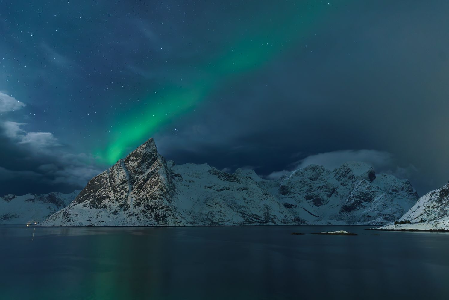 Aurora borealis glowing green over snow-capped mountains reflecting in calm Norwegian fjord waters at night.