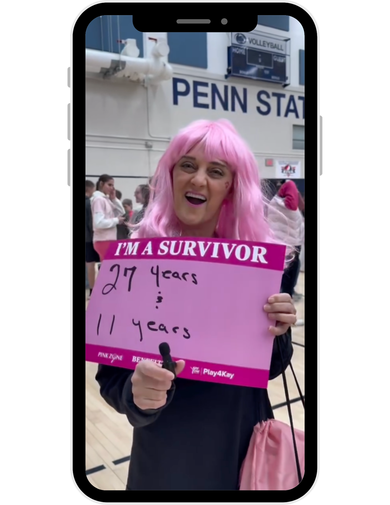 Penn State breast cancer survivor in pink wig holds I'm A Survivor sign showing 27 and 11 years at volleyball event.