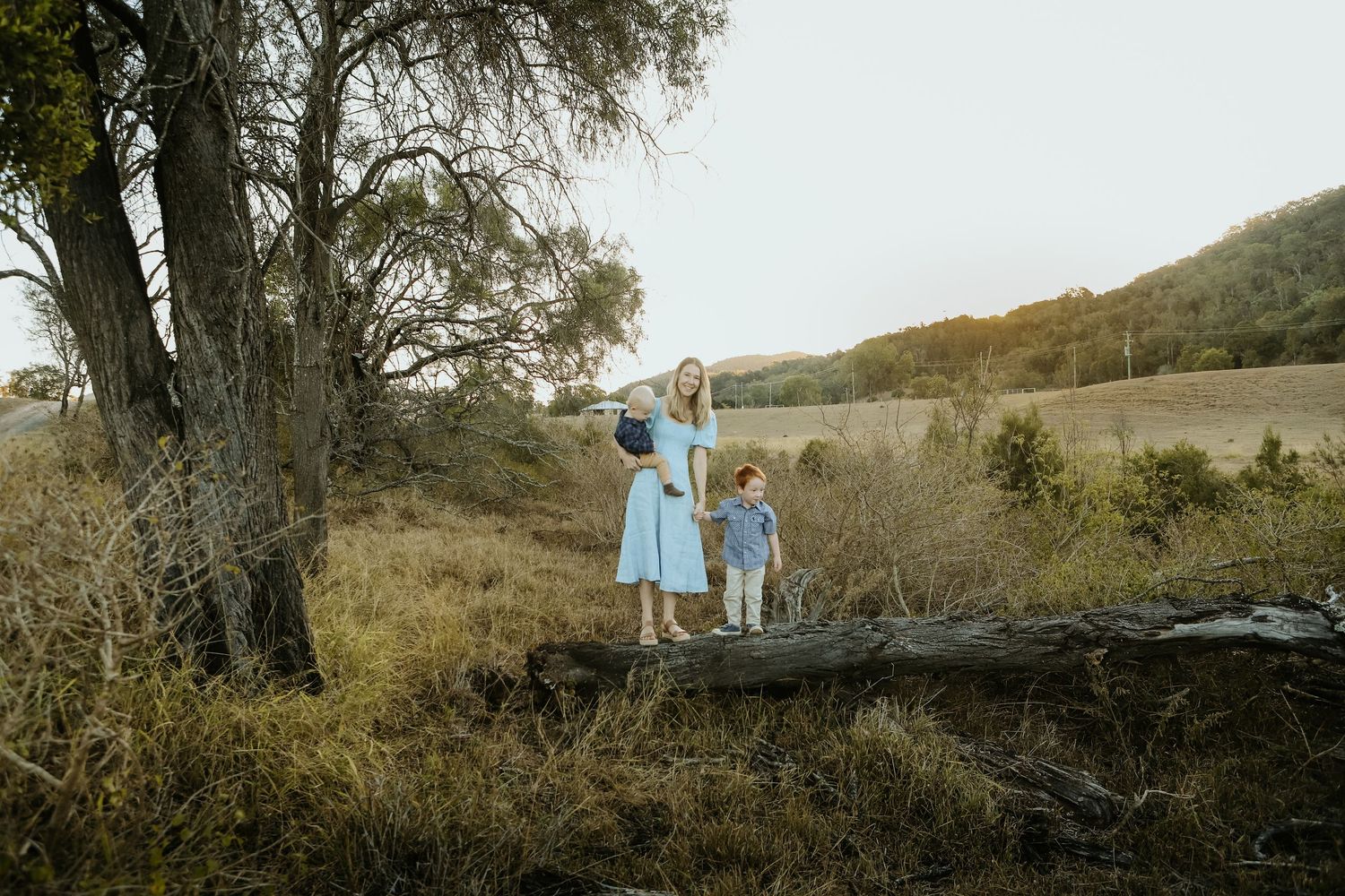 A mother and two children in matching blue outfits stand on a log in a scenic rural landscape with trees and hills.