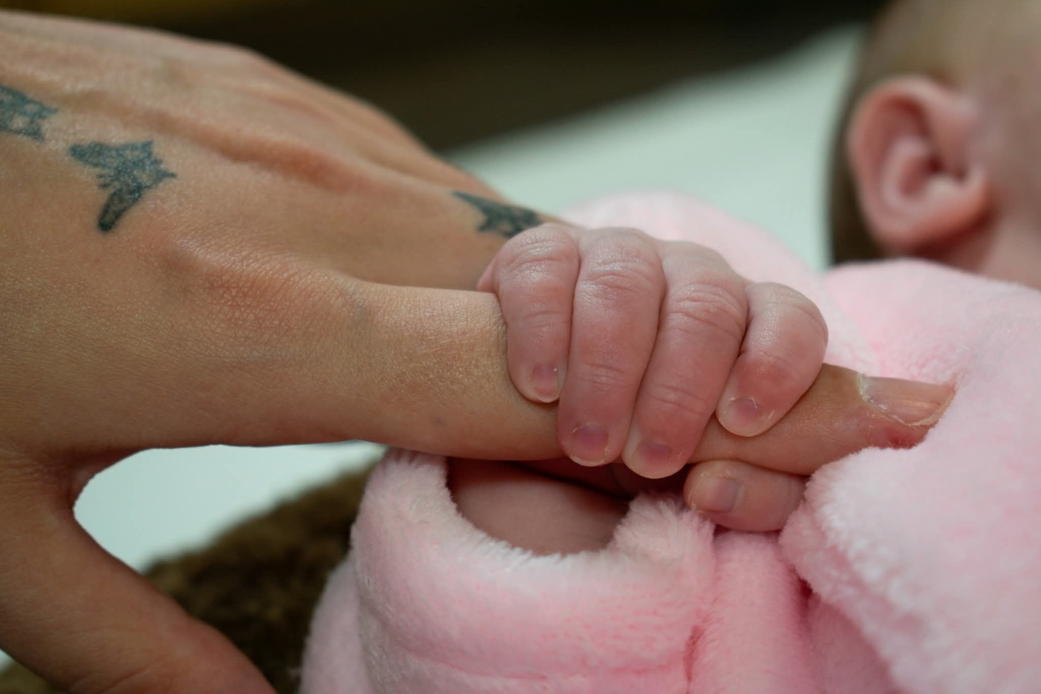 Close-up of a newborn baby gripping an adult finger with a butterfly tattoo on pink blanket.