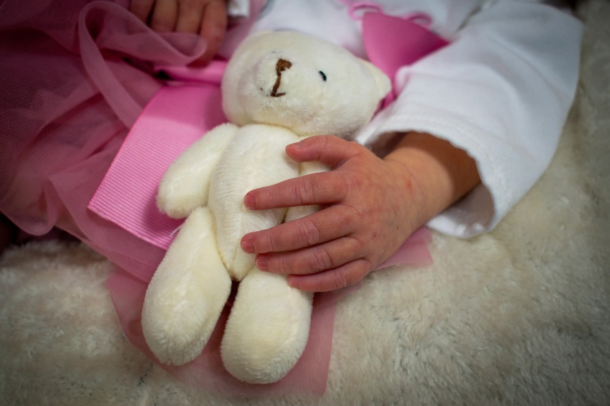 Baby hand gently holding a small white teddy bear on a soft white fur blanket.