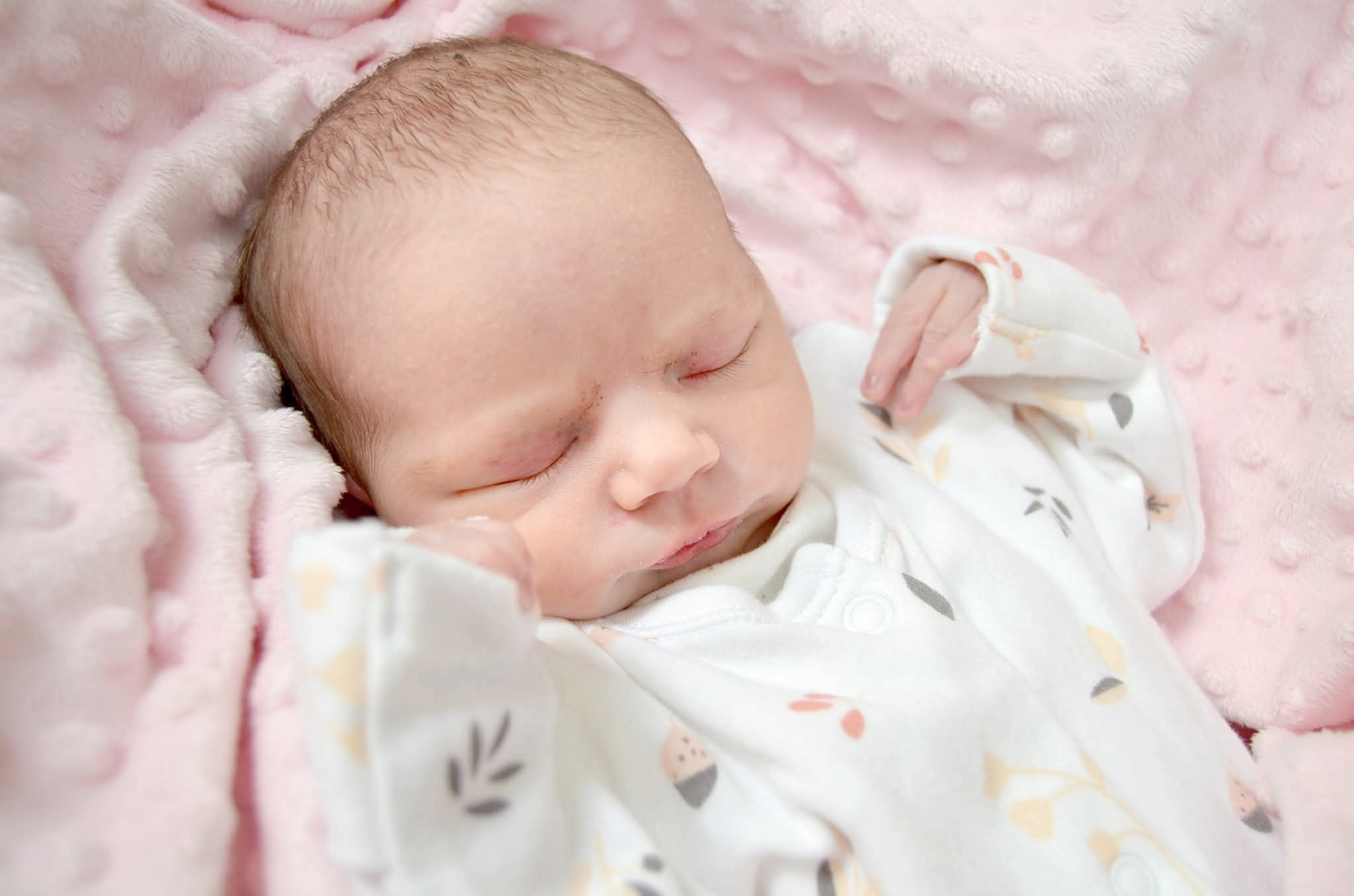 Peaceful newborn baby sleeping swaddled in a white floral blanket on a pink fluffy blanket.