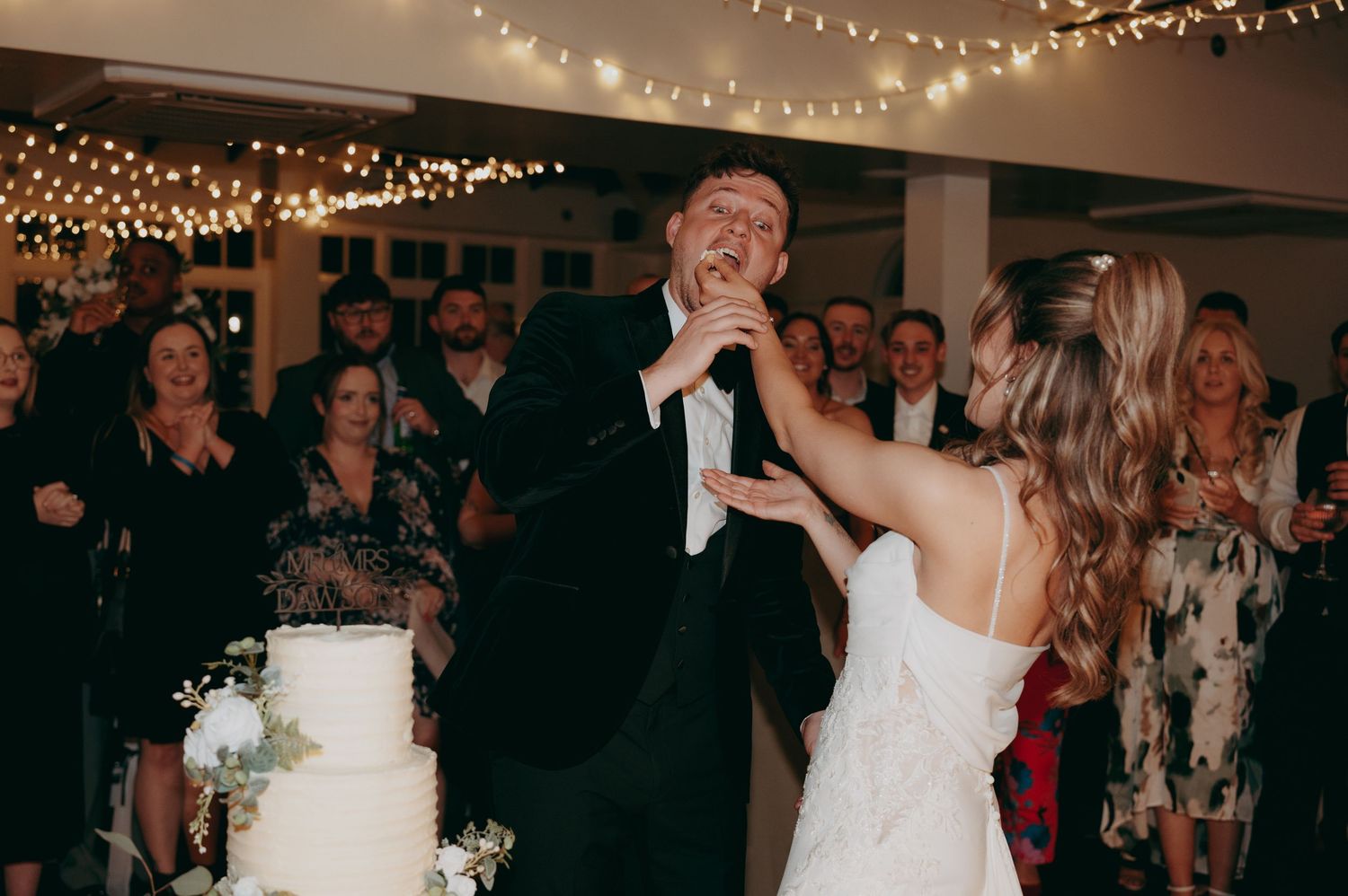 Bride and groom cutting wedding cake together while guests celebrate in background with fairy lights.